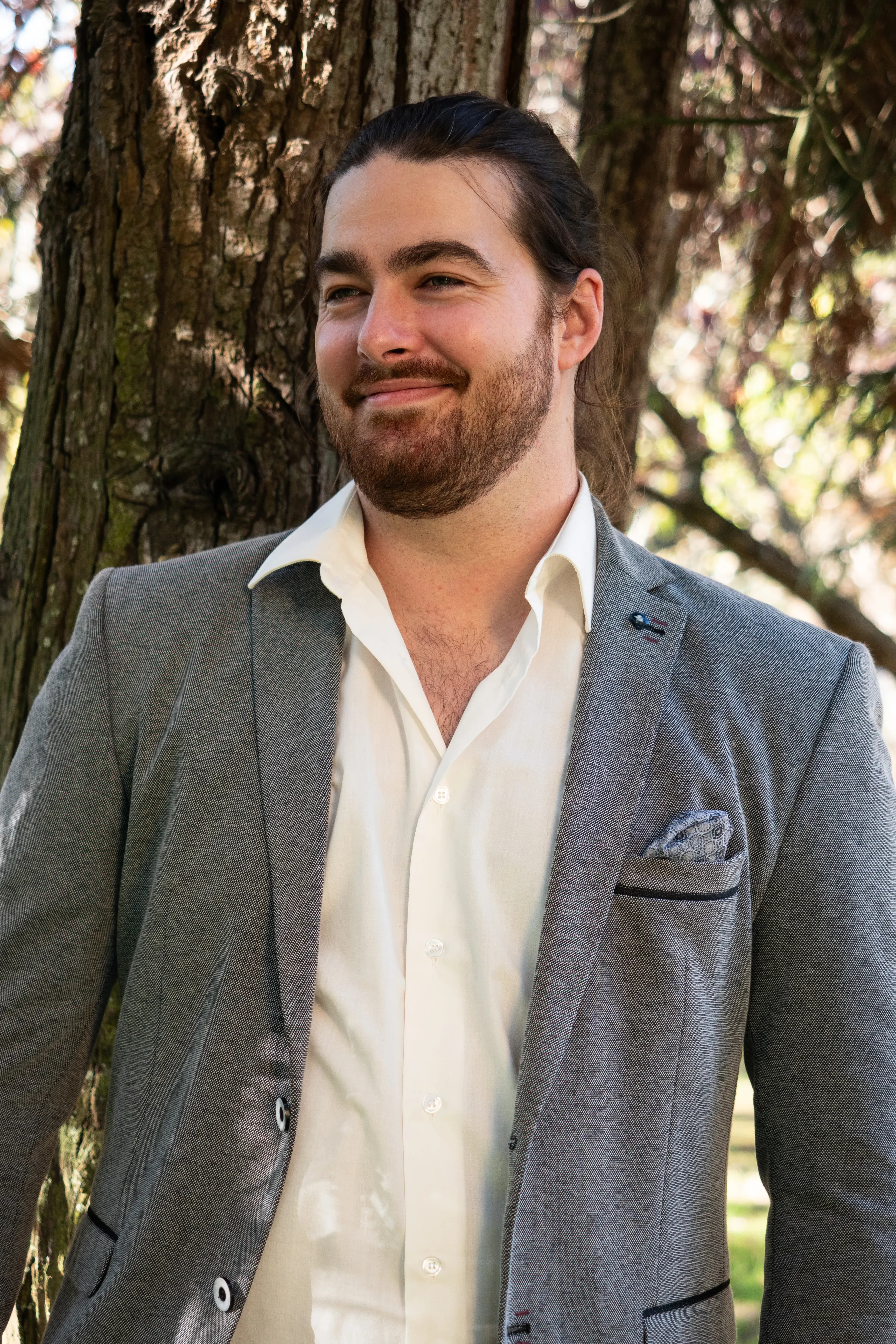 Retrato de um rapaz sorridente vestido com um blazer cinzento e uma camisa branca. Tem cabelo grande apanhado e uma barba aparada. Ao fundo tem uma árvore.